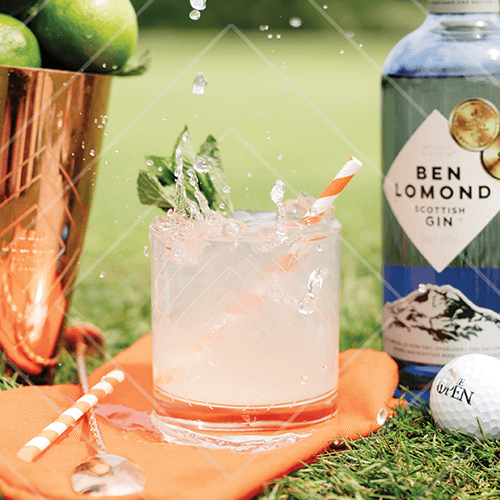 Refreshing pale cocktail splashing in a glass on grass beside a Ben Lomond gin bottle, with citrus garnish and striped straw on a sunny day.