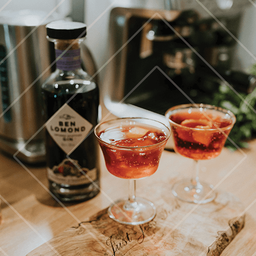 Two red spritz cocktails in coupe glasses on a wooden board with a Ben Lomond gin bottle in a kitchen setting.