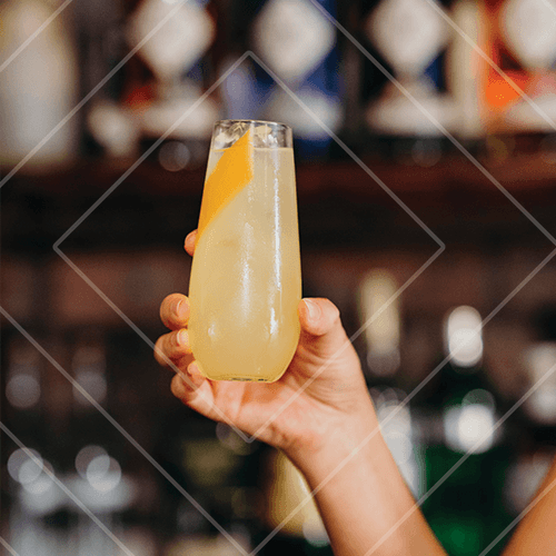 Hand holding a tall pale yellow cocktail with a citrus peel garnish in a softly lit bar environment.