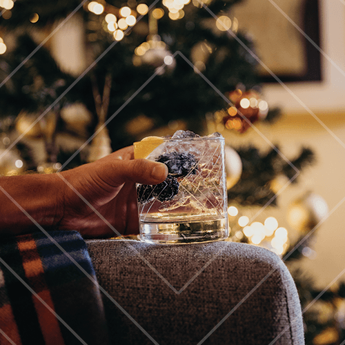 Hand holding a festive cocktail with berries and citrus in front of a softly lit Christmas tree.