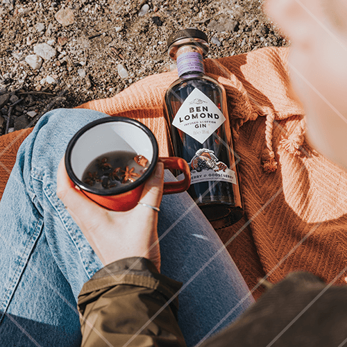 Person relaxing outdoors holding a warm spiced drink in a mug next to a bottle of Ben Lomond gin on a blanket.