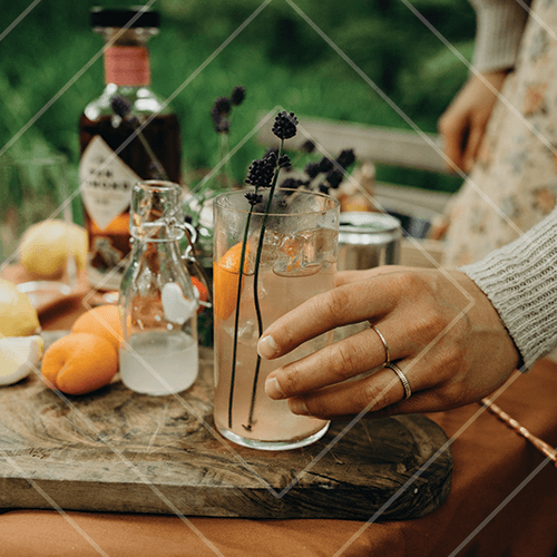 Hand holding a tall floral cocktail with lavender sprigs on a rustic outdoor table with citrus and ingredients.