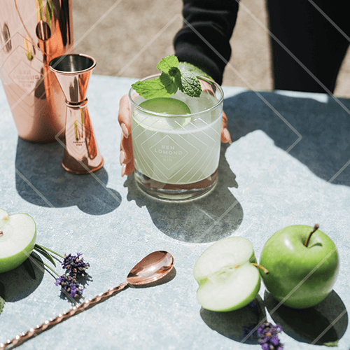 Fresh green cocktail with mint garnish on a bar surface surrounded by apples and copper bar tools.