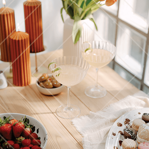 Two pale cocktails in coupe glasses with lime garnish on a sunlit table with strawberries, pastries, and candles.
