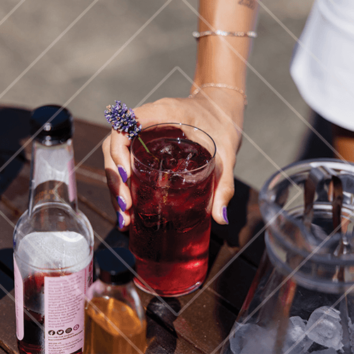 Close-up of a hand holding a deep red cocktail with ice and floral garnish on an outdoor table with bar tools.