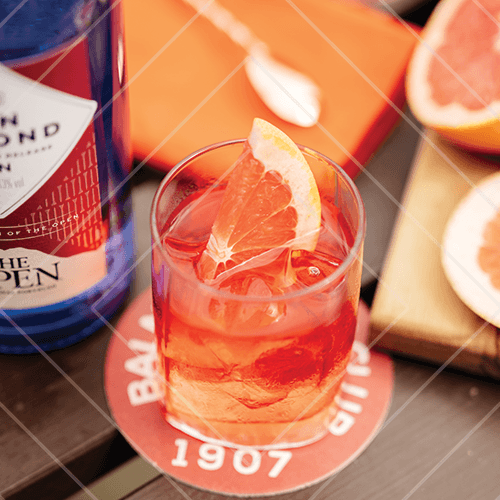 Bright red cocktail with grapefruit slice served on a coaster beside a blue Ben Lomond gin bottle.
