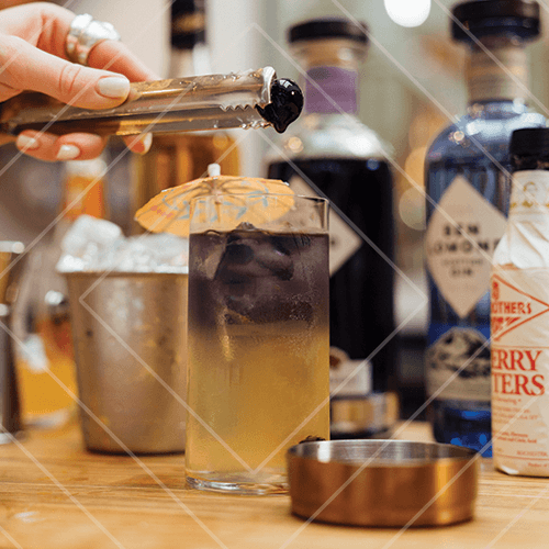 Layered purple and yellow cocktail with ice and umbrella garnish being finished with fruit in a bar setting.