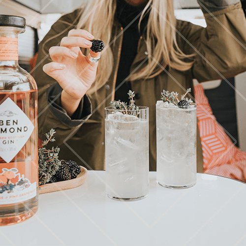 Bartender garnishing two tall iced cocktails with blackberries and herbs beside a bottle of pink gin.