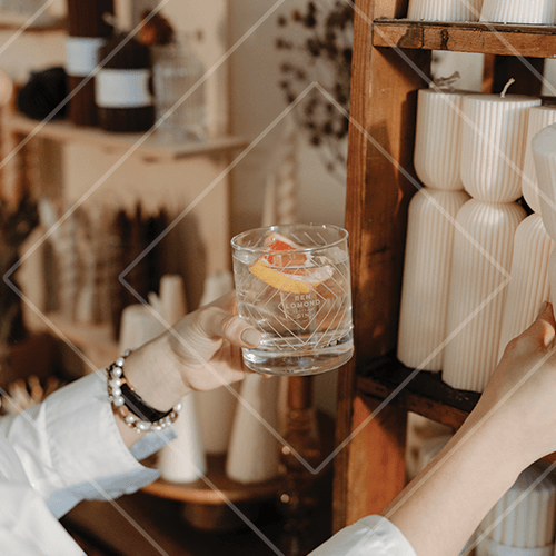 Hand holding a clear gin cocktail with ice and citrus peel beside a wooden shelf of sculptural ceramic vases in a warm interior.