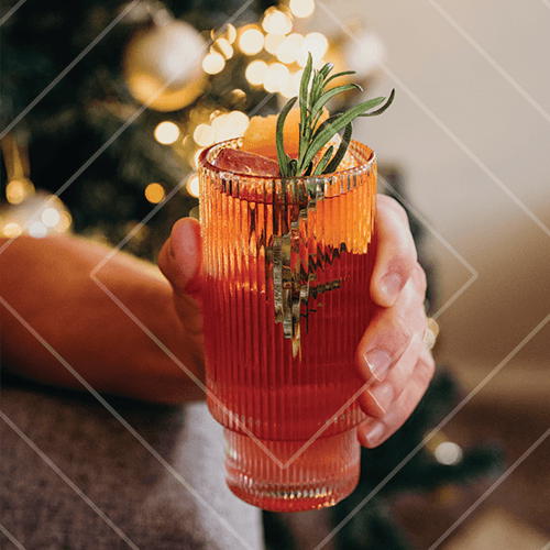 Warm-toned cocktail in a ribbed glass with rosemary garnish held in front of glowing holiday lights.