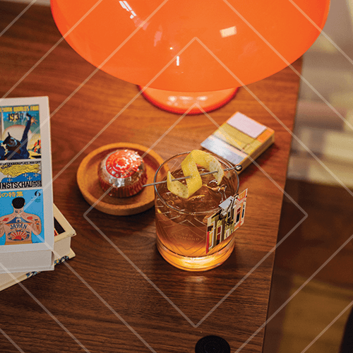 Cocktail with citrus peel and garnish on a wooden side table beside books and a glowing orange lamp.