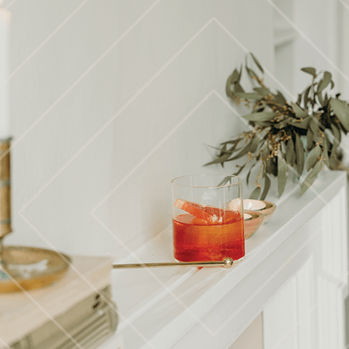 Amber cocktail in a tumbler on a mantelpiece with greenery and candles in a warm, minimalist interior.