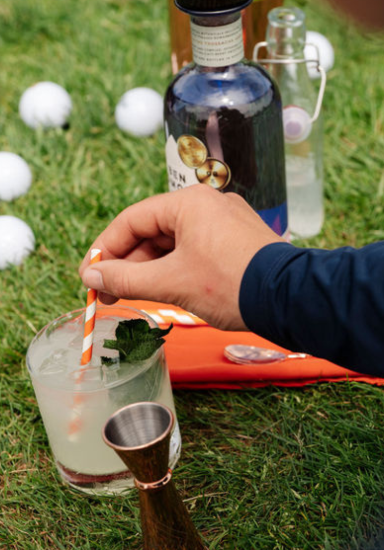 Person holding a cocktail with a straw on grass, with bottles and golf balls in the background