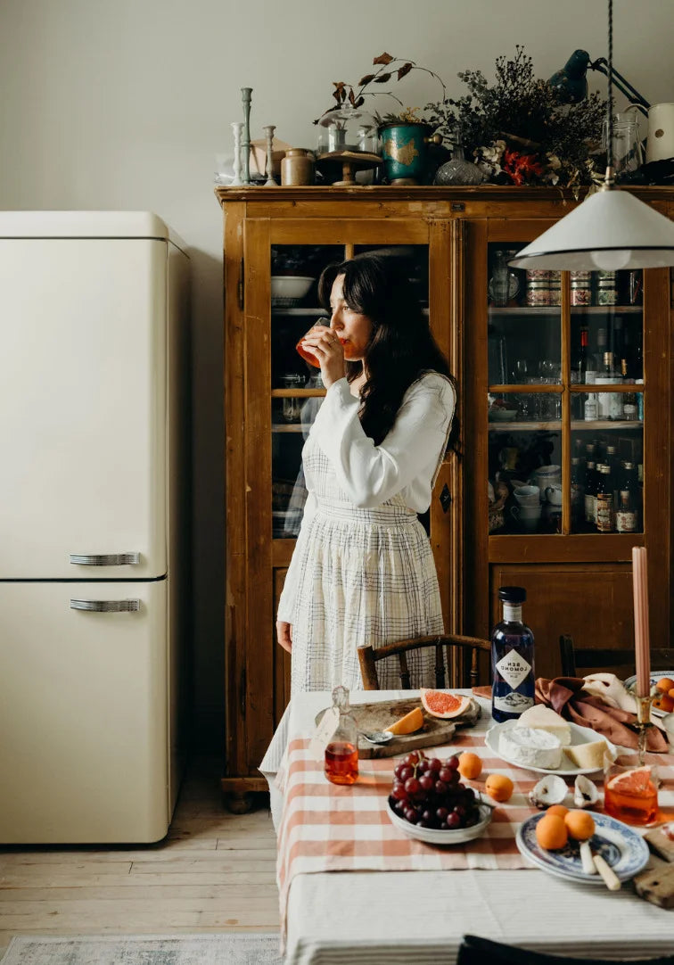 Woman standing in a kitchen with a table set for a meal, vintage refrigerator on the left.