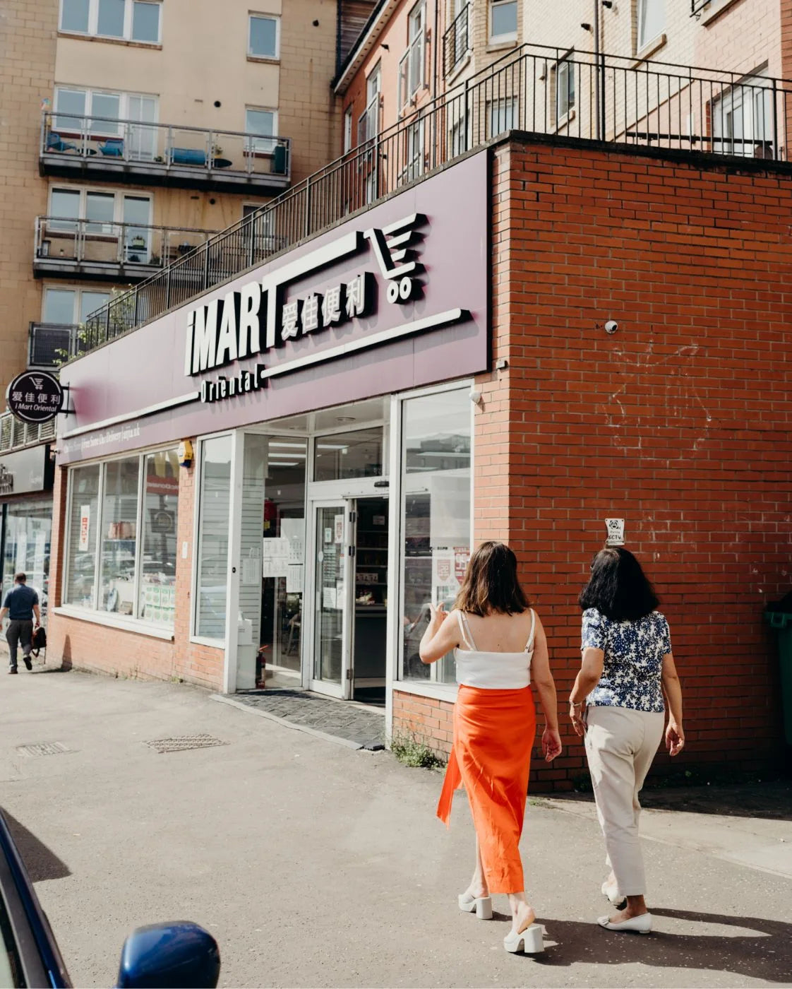 Two women walking past a store with a visible logo on a city street.