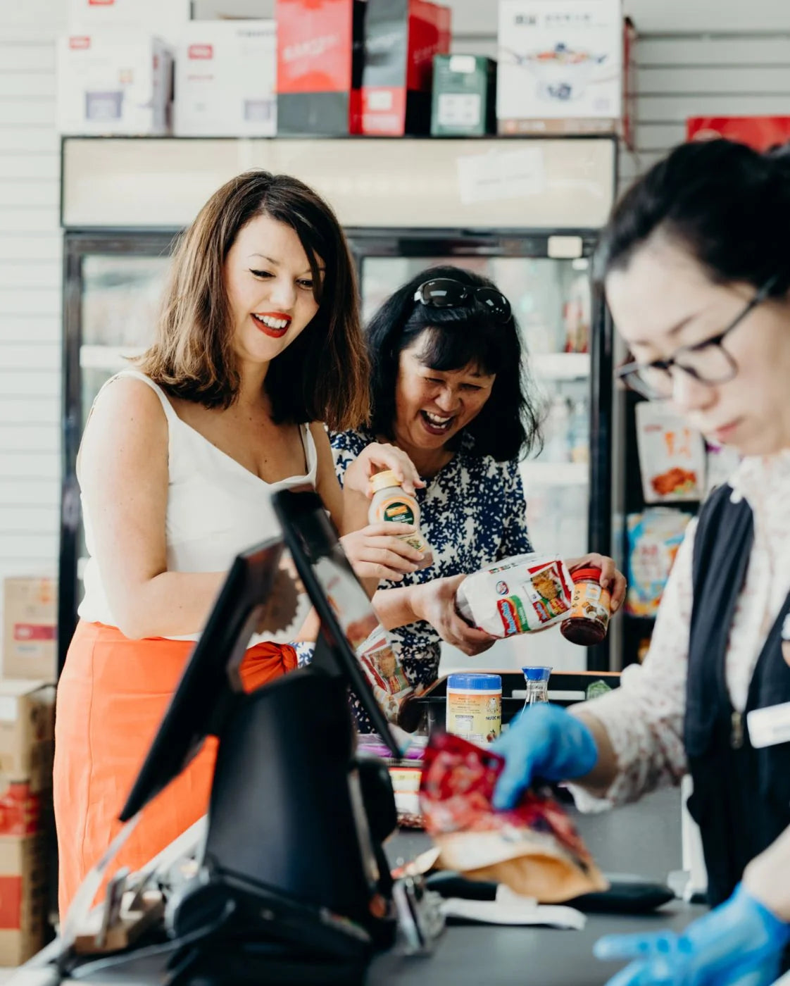 Three women in a store, two of whom are shopping and one is working at a checkout counter.