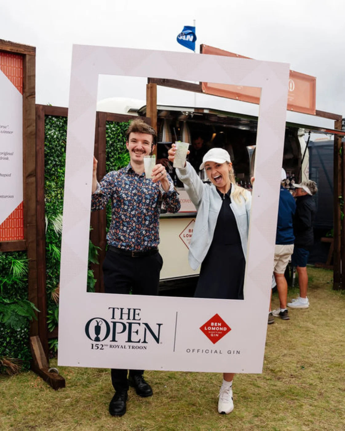 Two people posing inside a 'The Open' photo frame with a bar and outdoor setting in the background.