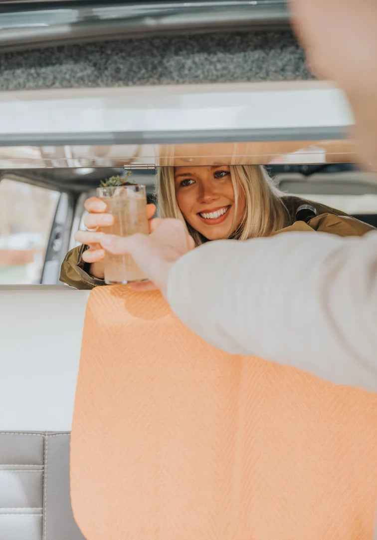 Woman holding a cocktail inside a parked campervan, smiling.