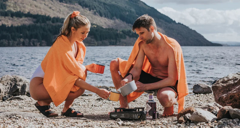 Two people cooking outdoors by a lake, wrapped in orange towels.