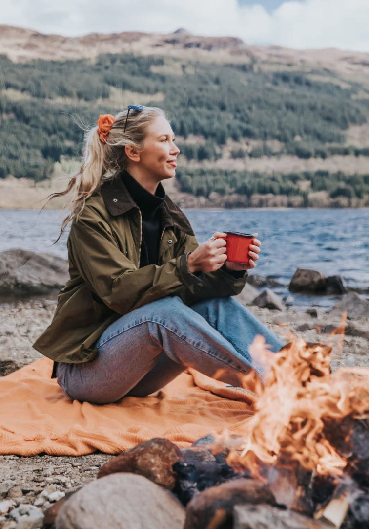 Person sitting by a campfire by a lake, holding a red mug