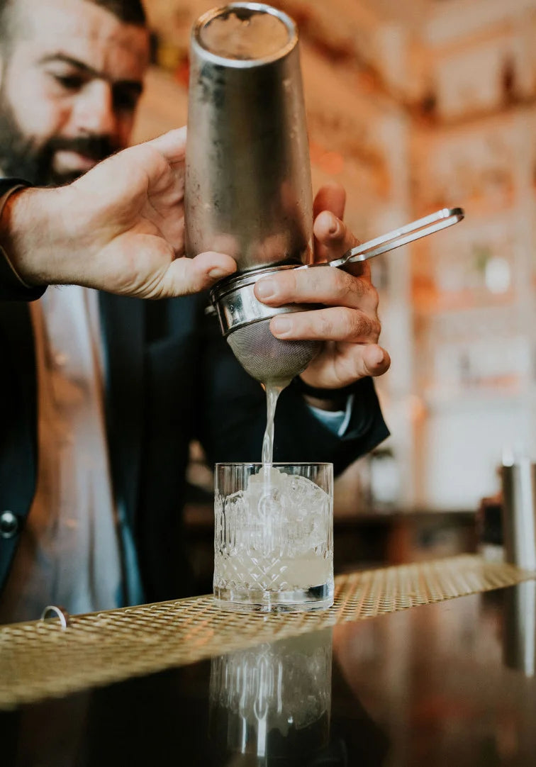 Person pouring a drink from a cocktail shaker into a glass with ice.