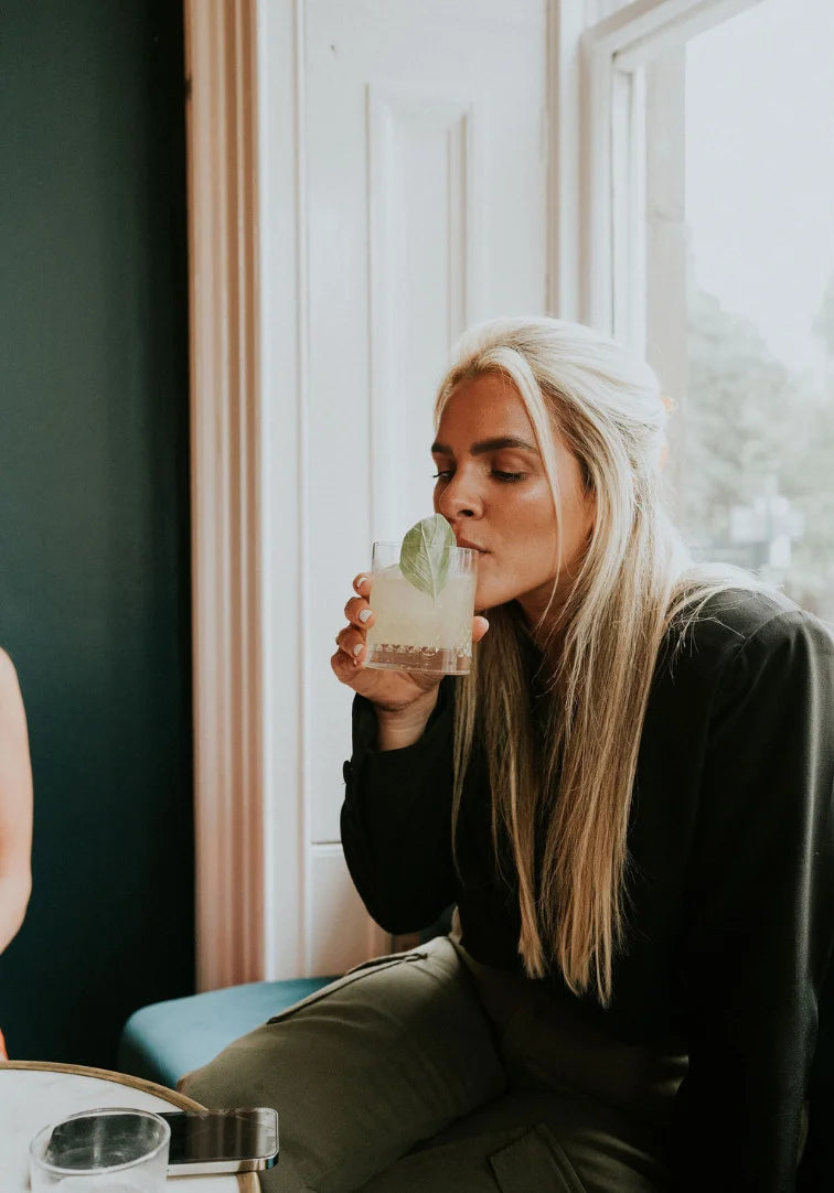Woman sitting indoors, holding a glass of a drink with a mint leaf.