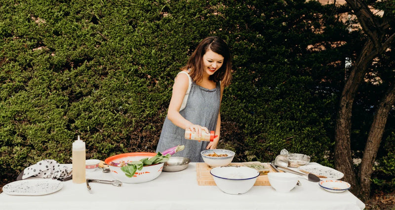 Woman preparing food outdoors at a table with greenery in the background