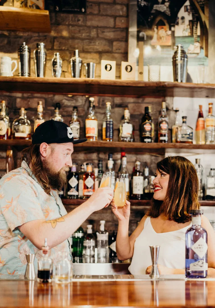 Two people at a bar clinking glasses with drinks in front of a well-stocked bar.