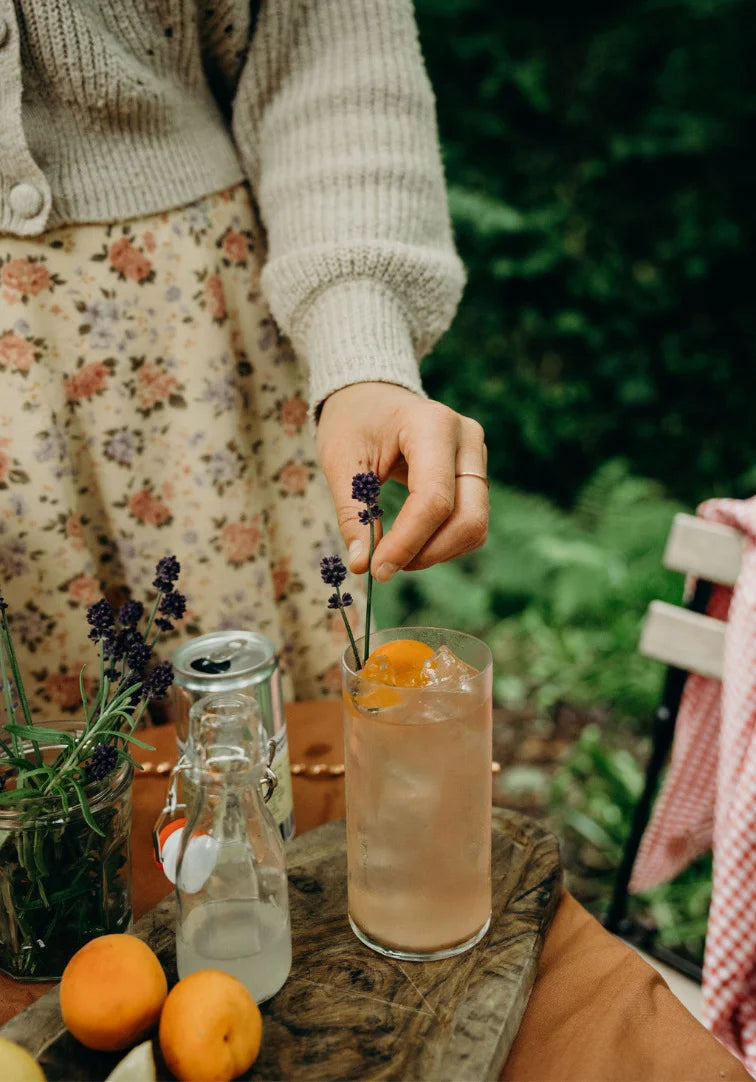 Person garnishing a cocktail with lavender and an orange on a wooden table outdoors.