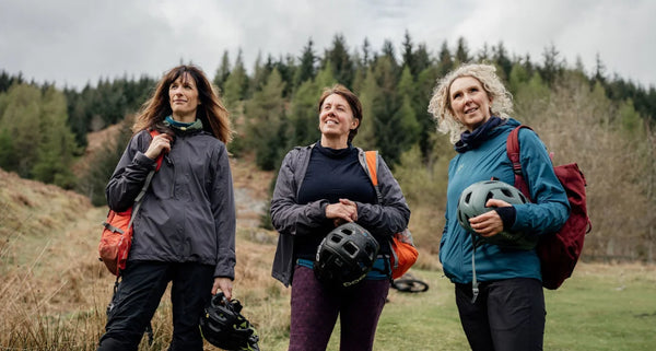 Three women with helmets and backpacks standing outdoors in a forested area.