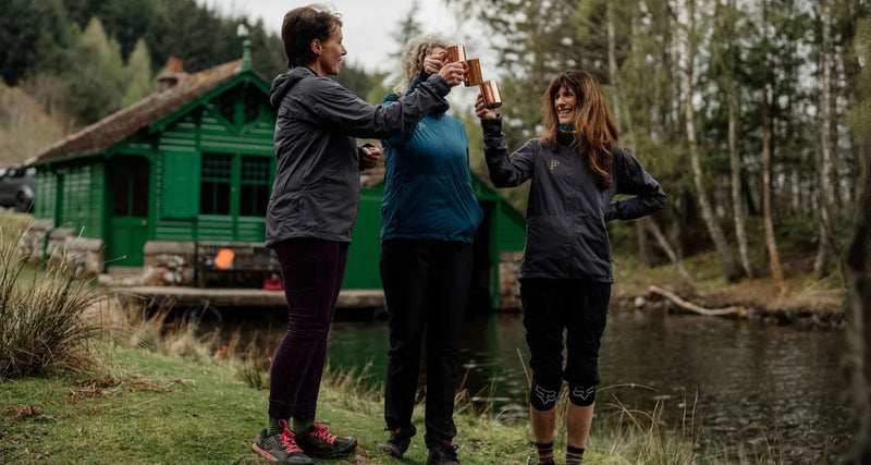 Three people standing by a lake, toasting with drinks, with a green cabin in the background.