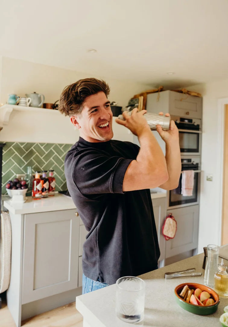 Man in a kitchen holding a cocktail shaker, smiling.