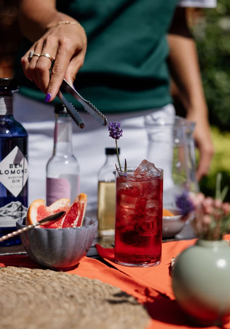 Cocktail being prepared with a red drink, garnished with a purple flower, on an outdoor setting.