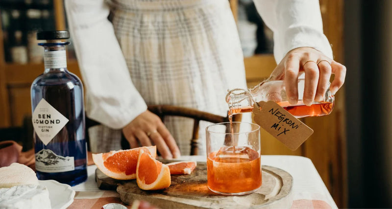 Person pouring a drink into a glass with a bottle of gin and grapefruit on a table.