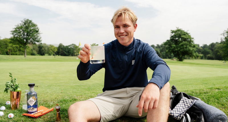 Man sitting on a golf course holding a glass of milk, with golf equipment and a bottle in the background.