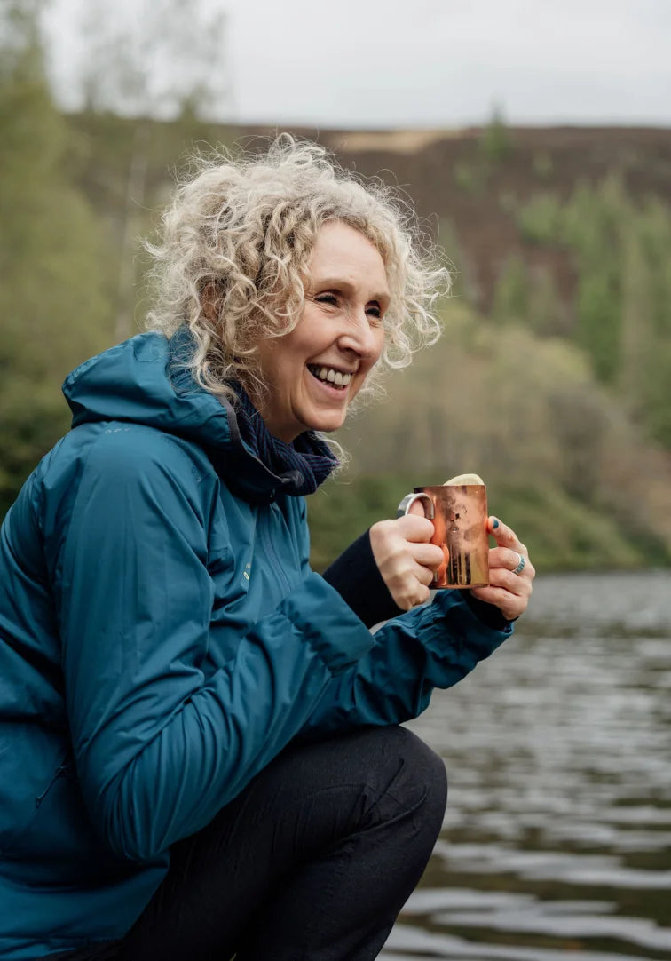 Woman in a blue jacket holding a mug by a lake