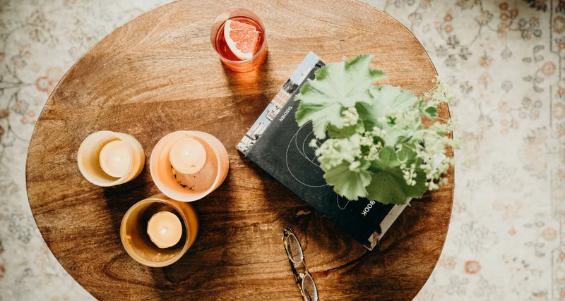 Wooden table with candles, a glass of orange juice, and a book on a patterned rug.