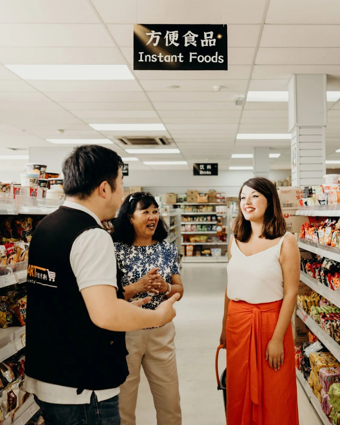 Three people in a convenience store with 'Instant Foods' sign above.