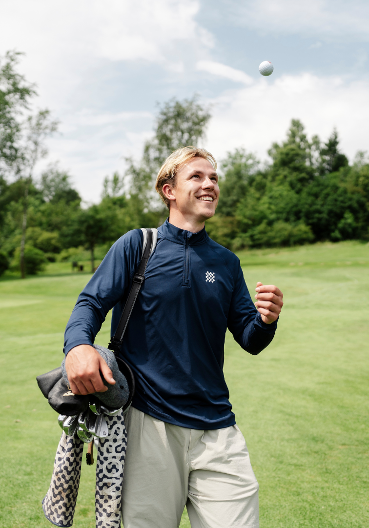 Man on a golf course holding a golf club and ball, wearing a navy blue jacket with a logo.