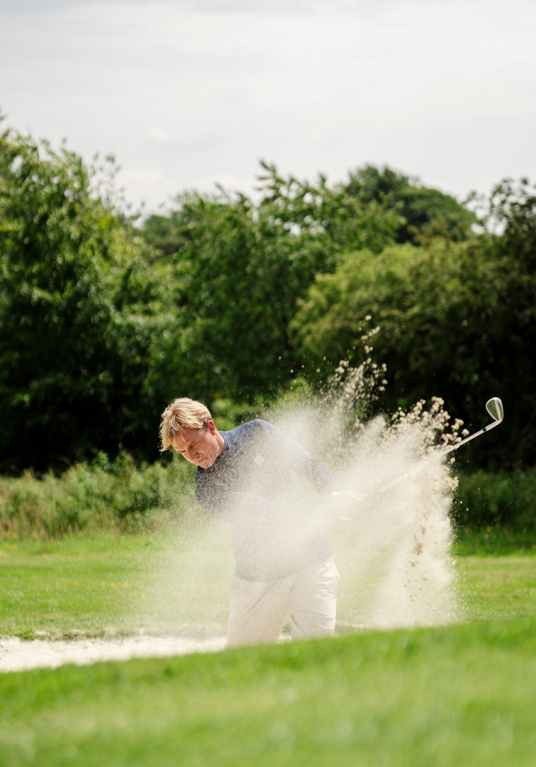 Man playing golf hitting a ball out of a sand trap with trees in the background