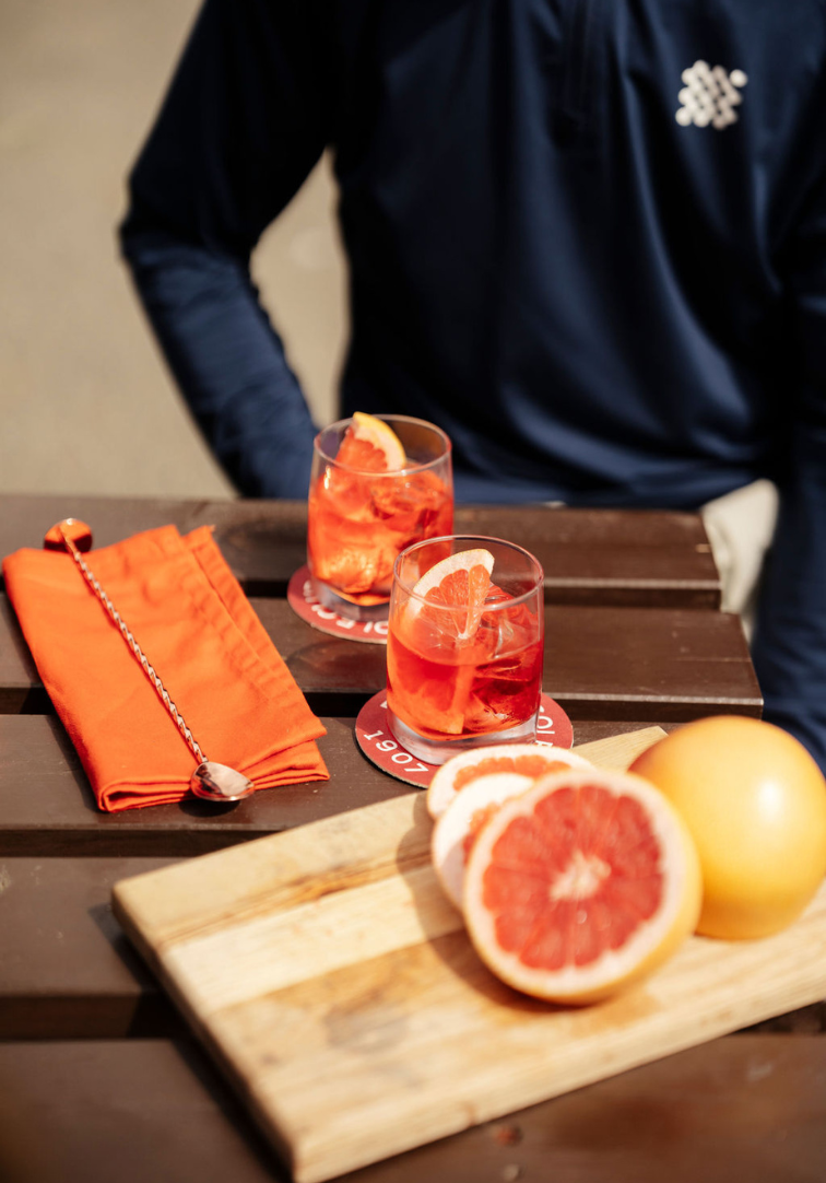 Two glasses of red cocktails with orange slices on a wooden table, accompanied by a cutting board with grapefruits.