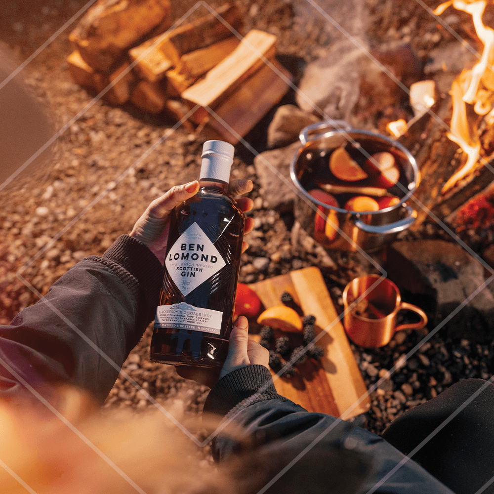 Person holding a bottle of Ben Lomond Scottish Gin by a campfire with hot cocktail and fruit.