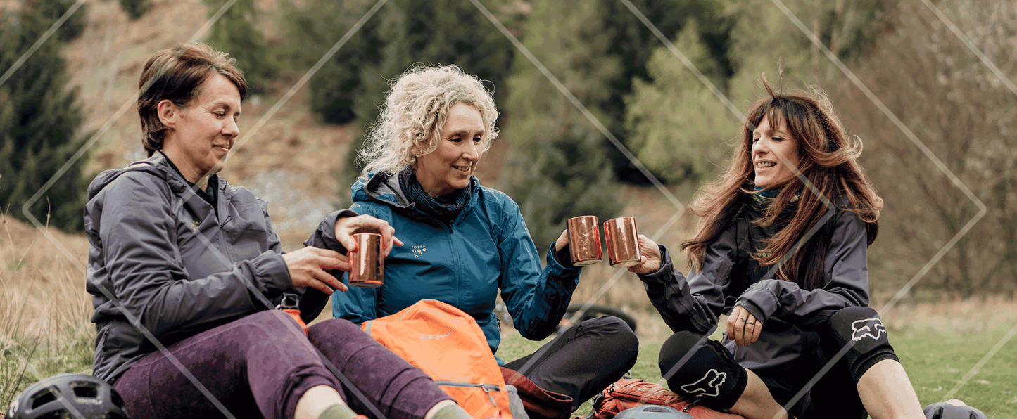 Three women sitting outdoors, possibly on a camping trip, with one holding a can.
