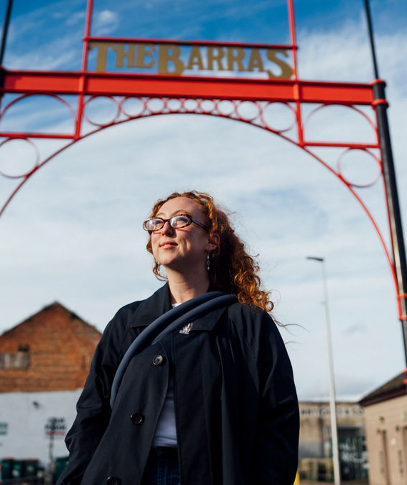 Person standing in front of 'The Barras' sign with a red archway.