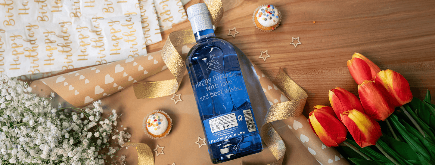 Blue bottle of gin with flowers, cards, and cupcakes on a wooden surface