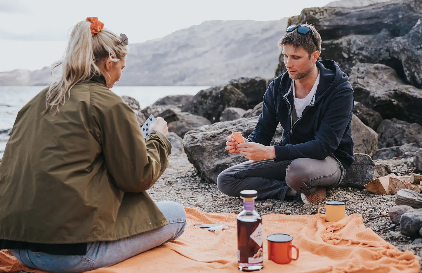 Two people sitting on a blanket by a body of water with drinks.