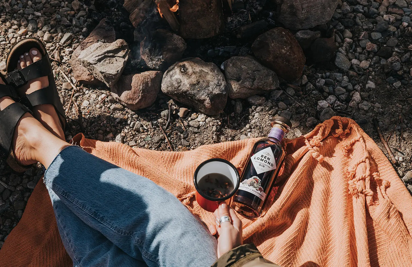 Person sitting on a rock with a bottle of beer and a cup, surrounded by nature.