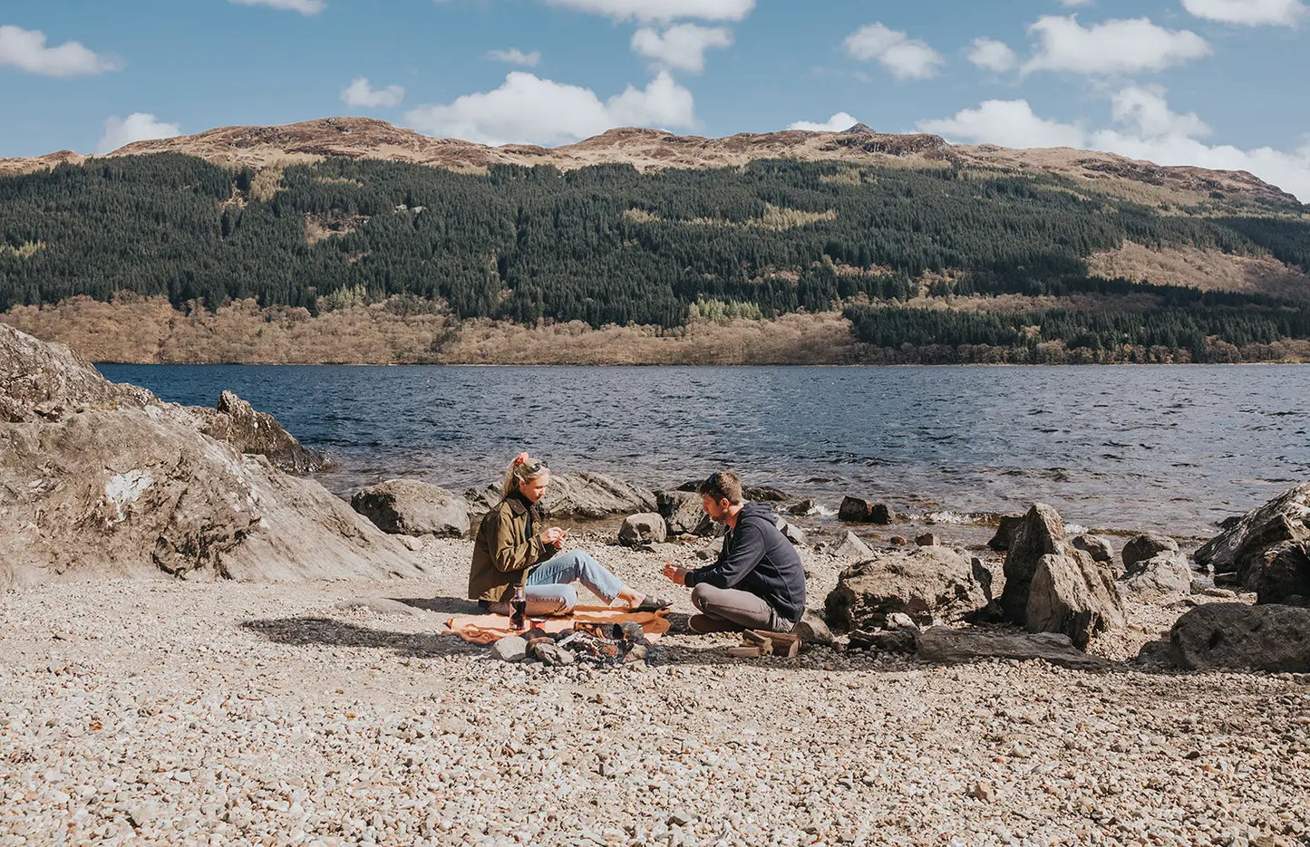 Two people sitting on a rocky beach with mountains and water in the background