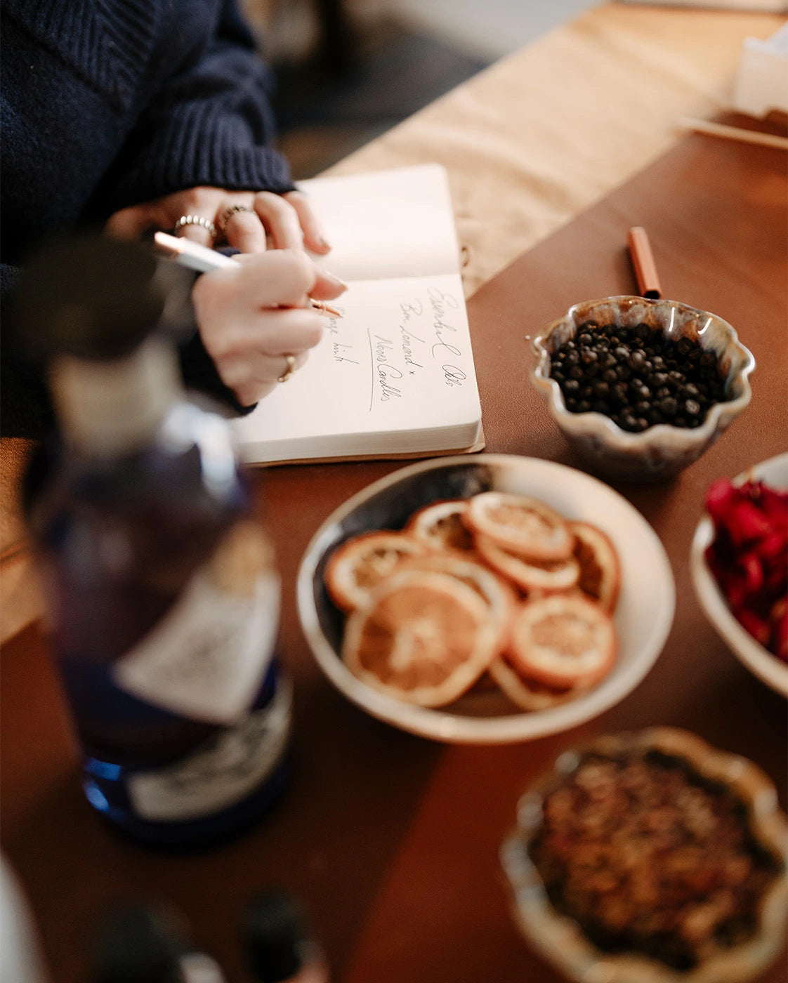 Person writing in a notebook with a bottle and bowls of dried fruit on a wooden table.