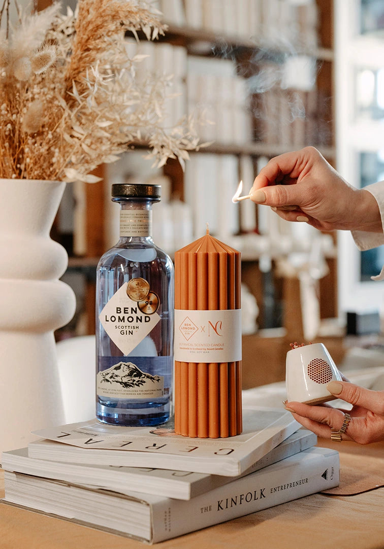 Person lighting a candle next to a bottle of gin on a table with decor items.